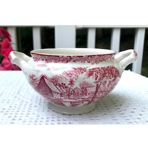 May include: A white ceramic creamer with two handles and a red and white floral pattern. The pattern features a cottage scene with trees and a river.