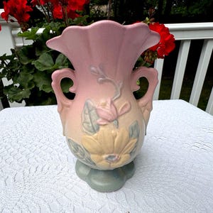 Puede incluir: Un jarrón de cerámica rosa con la parte superior con volantes y dos asas. El jarrón presenta un diseño floral con una flor amarilla y detalles en rosa y azul. La base es de color verde claro.