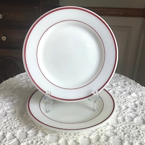 Puede incluir: Dos platos de cena de cerámica blanca con una sola banda roja alrededor del borde. Los platos están apilados uno encima del otro.