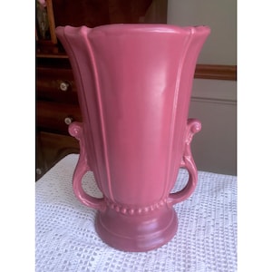 Puede incluir: Un jarrón de cerámica rosa vintage con dos asas y un diseño acanalado. El jarrón tiene un borde de cuentas alrededor de la parte superior e inferior.