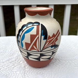 May include: A hand-painted ceramic vase with a cream-colored body, featuring geometric designs in black, blue, and rust-red. The vase has a terracotta-colored base and rim, and a scalloped edge. The vase is likely used for decorative purposes.