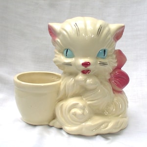 May include: A white ceramic planter shaped like a cat with a pink bow. The cat has blue eyes and a red mouth. The planter has a small pot for planting.