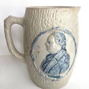 Puede incluir: Una jarra de gres vintage con una superficie texturizada y un asa. La jarra presenta un retrato azul y blanco de un hombre en un marco decorativo. El color general es beige claro.