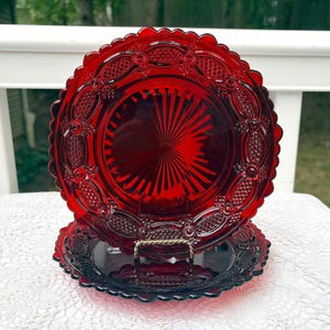 May include: Two circular ruby red glass plates featuring a pressed pattern with a central sunburst design, surrounded by an intricate border of diamond-shaped textures and oval motifs, finished with a scalloped edge along the outer rim.