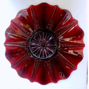 Può includere: Una grande ciotola da portata in vetro rosso scuro con un bordo a festoni e un motivo decorativo in rilievo al centro. La ciotola è in vetro rosso rubino e ha un aspetto vintage.