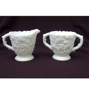 Könnte beinhalten: Ein weißes Milchglas-Sahnegießer- und Zuckergefäß-Set mit einem Blattdesign. Der Sahnegießer hat einen Griff und einen Ausguss. Die Zuckerdose hat zwei Griffe.