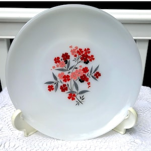 Puede incluir: Plato de cerámica blanco con diseño floral. El plato presenta un grupo de flores rojas, rosas y negras con hojas grises. El plato se exhibe sobre un soporte blanco, mostrando una estética vintage.