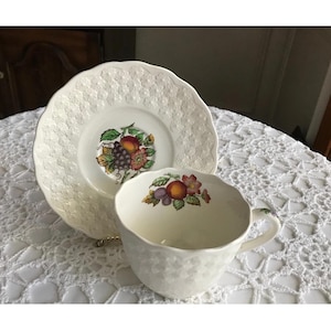 May include: A white teacup and saucer with a floral design featuring a cluster of fruit, including grapes, apples, and plums. The saucer has a raised, embossed pattern.