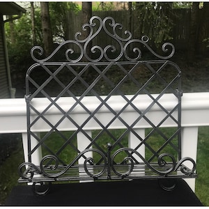 May include: A black metal decorative rack with a lattice design and scrolled accents. The rack has a shelf at the bottom and a decorative top with swirling details. The rack is designed to hold items and has a vintage aesthetic.