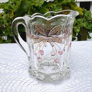 May include: A clear glass jug with a handle, adorned with gold-coloured leaves and pink cherries. The jug features a scalloped rim and a decorative band. A charming vintage piece, perfect for serving.