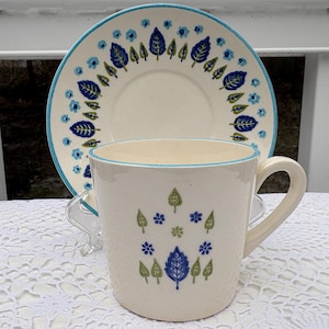 May include: Cream ceramic mug and saucer set with a textured finish. The mug and saucer are decorated with blue, green, and white leaf and floral patterns. The rim of the mug and saucer has a light blue border. A charming tea set.