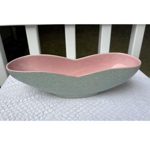 Op de afbeelding: Een keramische kom in mid-century modern stijl met een unieke vorm. De binnenkant is zachtroze, contrasterend met de gespikkelde, lichtgroene buitenkant. De kom heeft een langwerpige vorm, ideaal voor decoratie of serveren. Ongeveer 30 cm lang.