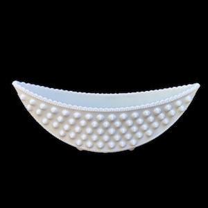 Puede incluir: Un cuenco decorativo blanco, de forma ovalada, con una superficie texturizada de pequeños puntos en relieve. El borde está detallado con un patrón de cuentas. El cuenco se apoya en tres pequeños pies, sobre fondo negro.