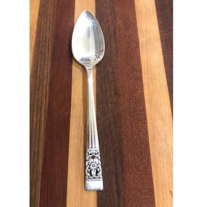 May include: A silver teaspoon with an ornate, floral design on the handle. The spoon is lying on a wooden surface.