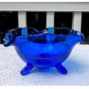 Puede incluir: Cuenco de vidrio azul cobalto con borde rizado y tres patas. El cuenco tiene una banda plateada alrededor del centro.