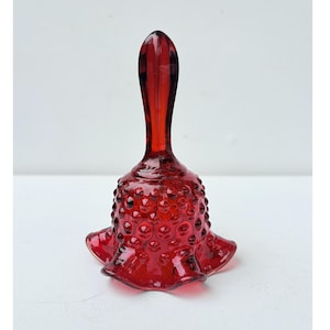 Puede incluir: Una campana de cristal rojo rubí translúcido con una textura de puntos en relieve en el cuerpo. La campana tiene un mango liso y alargado y un borde acampanado y ondulado en la base, creando una silueta distintiva con acabado brillante.