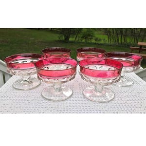 May include: Set of six clear glass dessert cups with a red rim. The cups have a textured design and are on a white lace tablecloth.