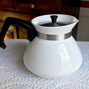 May include: A white glass coffee pot with a black handle and lid. The pot has a silver band around the middle. The pot is sitting on a white lace tablecloth. The pot is a vintage style.