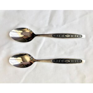 May include: Two silver spoons with black and gold decorative designs on the handles. The spoons have a rounded bowl shape.
