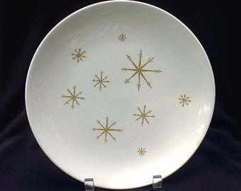 May include: A white ceramic plate with a mid-century modern design. The plate features a pattern of gold-colored snowflakes. The plate is round and has a slightly raised edge. The plate is likely used for serving food.