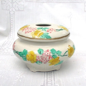 May include: A white ceramic jar with a lid, decorated with hand-painted yellow, pink, and green flowers. The jar has a round body and a slightly flared rim.