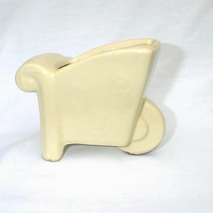 Puede incluir: Un pequeño macetero de cerámica en forma de carretilla de color crema. La carretilla tiene un asa redondeada y una sola rueda.