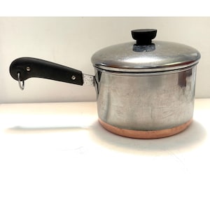May include: A stainless steel saucepan with a copper-colored base, featuring a black handle with a small metal hanging loop and a matching stainless steel lid topped with a black knob. The pot has a cylindrical body and a polished finish.