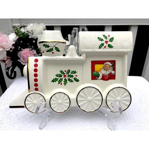 Puede incluir: Tren de Navidad de cerámica blanca con detalles de acebo rojo y verde. Santa Claus está en la ventana del último vagón. El tren tiene ribetes dorados y puntos rojos en el lateral.
