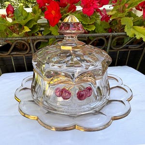 May include: A clear glass covered dish with a gold rim and a scalloped base. The dish features a cherry design and gold leaf accents. The lid has a decorative finial. The dish is sitting on a white surface.