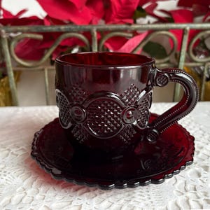 Puede incluir: Un juego de taza y platillo de color rojo rubí. La taza tiene un asa decorativa y un patrón detallado alrededor de la taza y el platillo. El juego está hecho de vidrio y se encuentra sobre un mantel de encaje blanco.