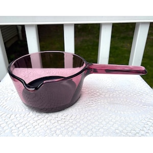 Puede incluir: Una cacerola vintage de vidrio transparente en un vibrante tono púrpura. La cacerola presenta un mango largo y resistente y un pico vertedor. Este artículo de cocina es ideal para cocinar y servir.