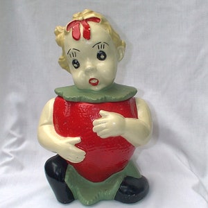 Puede incluir: Un tarro de galletas de cerámica vintage con forma de niña sosteniendo una fresa roja. La niña tiene el pelo rubio con un lazo rojo, ojos verdes y un cuello verde.