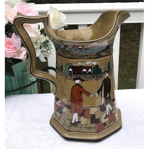 May include: A brown ceramic pitcher with a scene of two men shaking hands in front of a building. The pitcher has a black handle and a brown and black patterned base. The text "most cordial hospitality" is written on the base.
