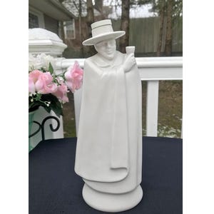 May include: White ceramic figurine of a person wearing a hat and robe, holding a small cup. The figurine is standing on a white base.