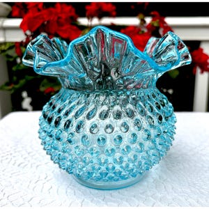 Könnte beinhalten: Eine hellblaue Hobnail-Glasvase mit gerüschtem Rand. Die Vase hat einen runden Körper mit einer strukturierten Oberfläche aus kleinen, erhabenen Noppen. Der gerüschte Rand verleiht der Vase einen dekorativen Touch.