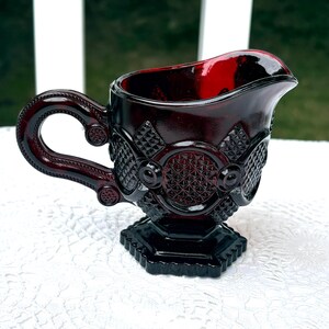 Peut inclure: Un pot à crème en verre rouge foncé présentant un motif en losanges pressés, un motif circulaire sur le côté et une anse robuste. Le récipient repose sur une base hexagonale et possède un bec verseur prononcé, affichant une riche couleur rubis.