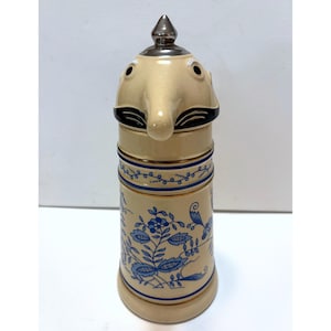 Peut inclure: Une chope à bière en céramique beige avec un couvercle en forme de visage et un dessus argenté. Le corps de la chope est décoré de motifs floraux bleus et d'une bordure bleue. La chope a une anse et une base.