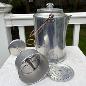 May include: A vintage aluminum coffee percolator with a glass knob on the lid. The percolator has a copper-colored handle and comes with its internal components, including a filter basket and a spreader. The components are arranged on a white surface.