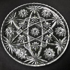 May include: A clear glass cake plate with a starburst design. The plate has a raised edge and a scalloped rim.