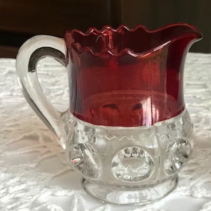 Puede incluir: Un pequeño creamer de vidrio transparente con una parte superior de vidrio rojo. El creamer tiene un borde festoneado y un asa de vidrio transparente. La parte inferior del creamer tiene un patrón de círculos en relieve.
