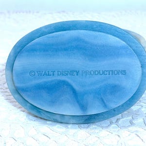 May include: An oval-shaped blue and white marbled base with a smooth, polished finish. The text "&copy; WALT DISNEY PRODUCTIONS" is clearly embossed in the center of the surface. The object features a swirling pattern of light and dark blue tones.