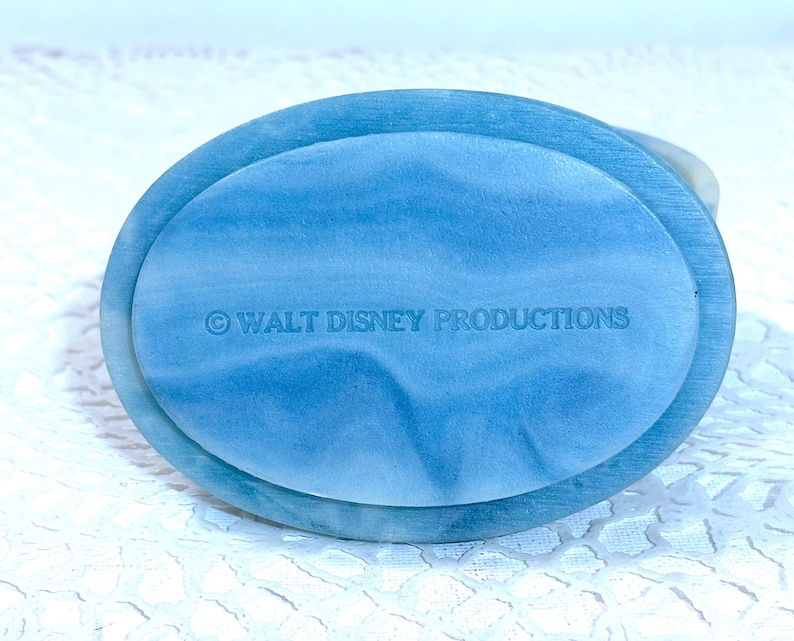 May include: An oval-shaped blue and white marbled base with a smooth, polished finish. The text "&copy; WALT DISNEY PRODUCTIONS" is clearly embossed in the center of the surface. The object features a swirling pattern of light and dark blue tones.