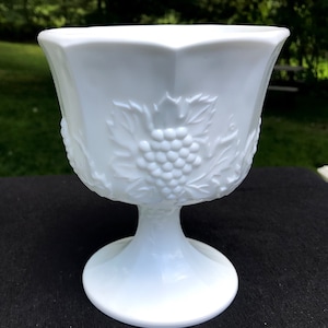 Puede incluir: Un tazón de pedestal de vidrio de leche blanco con un diseño de vid. El tazón es redondo y tiene una base ancha.