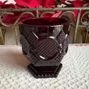 May include: A dark red glass goblet with a geometric pattern. The goblet has a square base and a detailed design around the body. The glass has a glossy finish and is set against a white lace tablecloth.