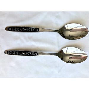 May include: Two silver spoons with black handles featuring a decorative design. The spoons have a shiny, reflective surface.