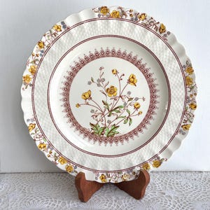 以下が含まれることがあります： 黄色の花と緑の葉のフローラルデザインが特徴の、スカラップエッジの白いセラミックプレート。プレートの縁には織り目加工が施され、マルーンのボーダーがあります。プレートは木製のスタンドに展示されています。
