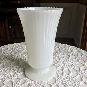 May include: A white milk glass vase with a fluted design and a scalloped rim. The vase has a pedestal base.