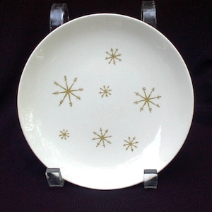 Op de afbeelding: Een witte keramische plaat met een modern design uit het midden van de eeuw. De plaat heeft een patroon van goudkleurige sneeuwvlokken. De plaat is rond en heeft een iets verhoogde rand. Een decoratief stuk voor het serveren van voedsel.