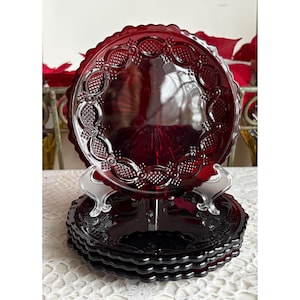 Puede incluir: Un juego de seis platos de vidrio rojo rubí con un borde decorativo festoneado. Los platos tienen un patrón floral y geométrico en relieve. Un plato se exhibe en un soporte plateado, mostrando el intrincado diseño y color.