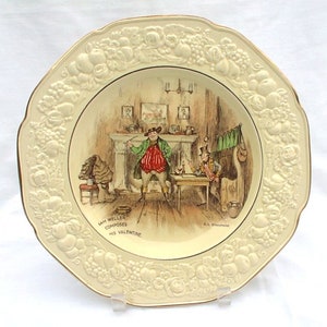 Può includere: Un piatto in ceramica color crema con un bordo floreale in rilievo. Il centro del piatto presenta un'illustrazione in bianco e nero di due uomini in una stanza. Il testo "Sam Weller Composes His Valentine" è stampato sotto l'illustrazione.